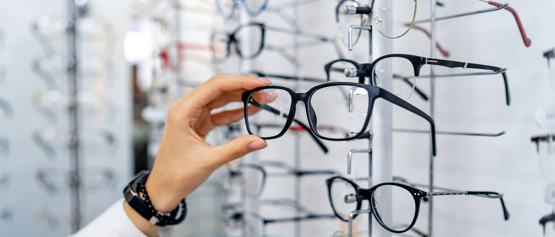 Eine Hand greift nach einer eleganten schwarzen Brille, die auf einem Ständer in einem Optikgeschäft ausgestellt ist, umgeben von verschiedenen anderen Brillengestellen.